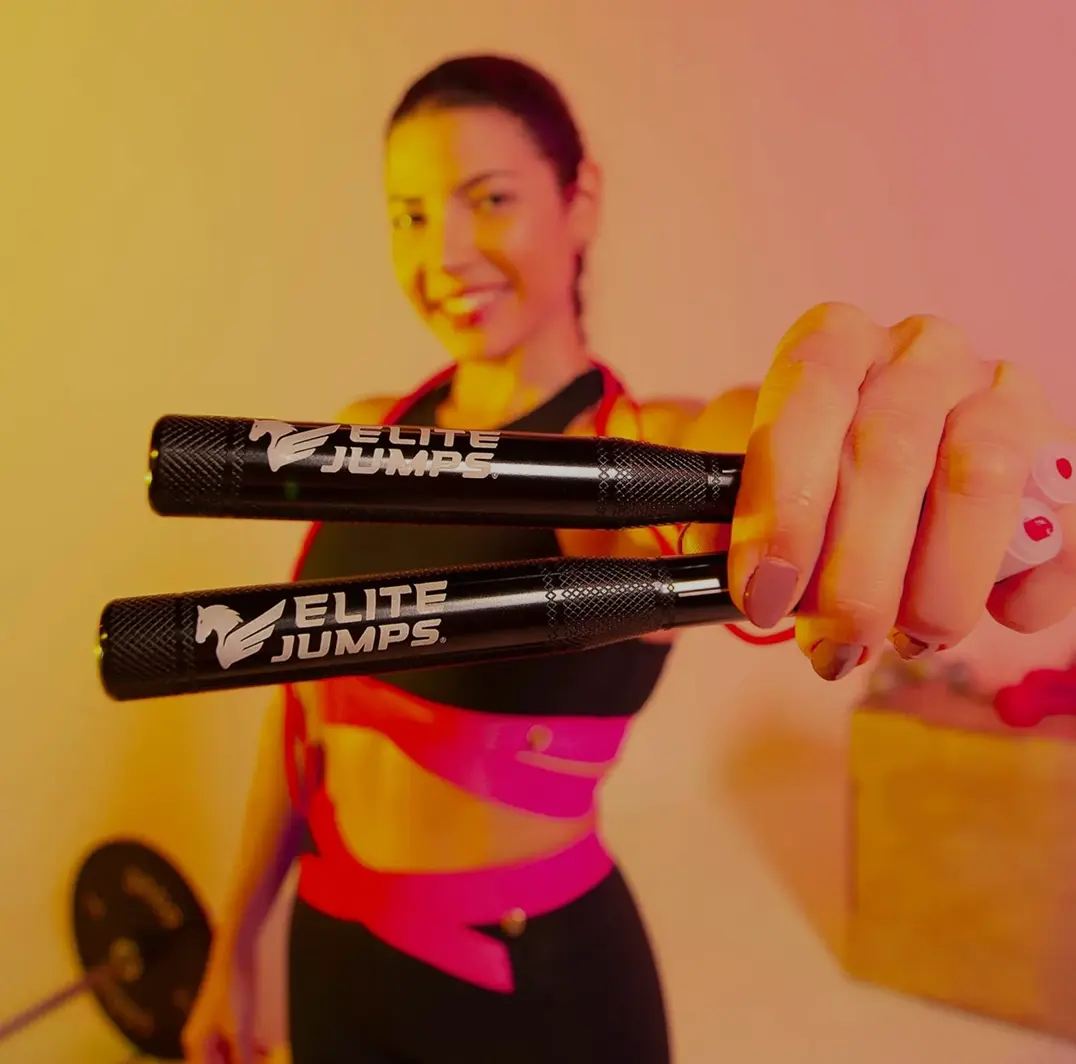 A woman in workout attire smiles while holding a pair of "Elite Jumps" jump rope handles toward the camera, with a gym setting and the "Elite Jumps" logo in the corner.