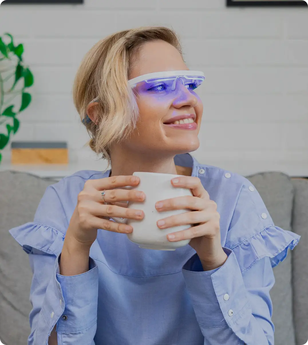 A smiling blonde woman wearing AYO light therapy glasses while holding a mug, relaxing on a couch at home