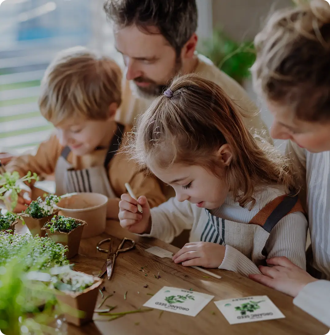 Father and three young children planting seeds together at a wooden table indoors, with a 'Spade to Fork' brand label in the corner.