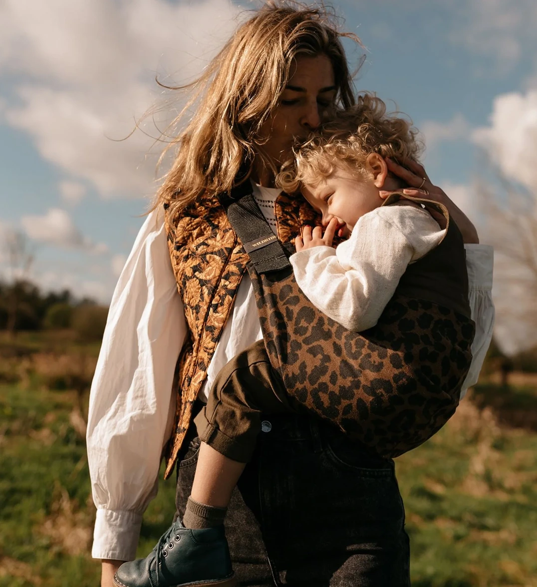 Mother in a field kissing her toddler, carried on her back in a leopard-print Wildride baby carrier, with a 'Wildride' brand label in the corner