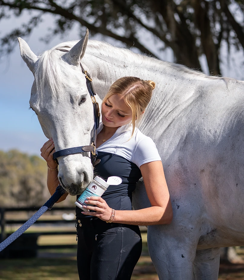 Woman hugging white horse while holding equine supplement bottle