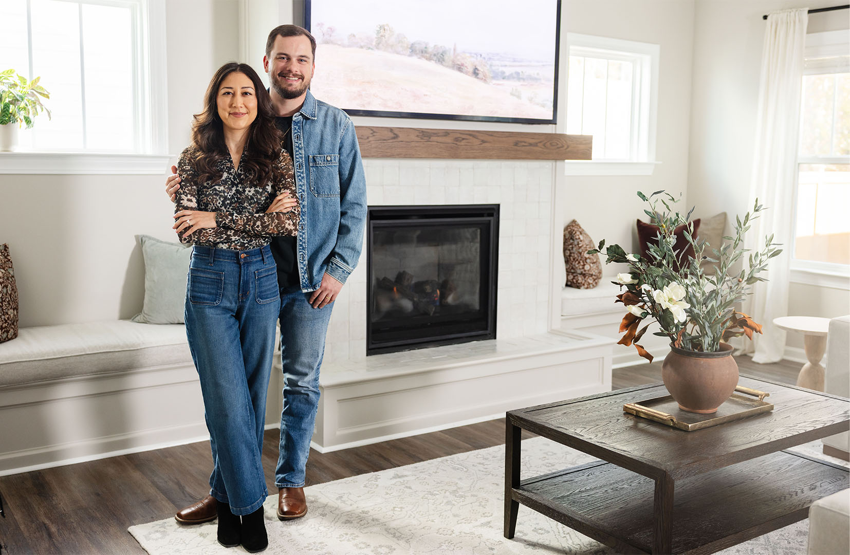 Smiling couple standing in a modern living room with a fireplace and a wooden coffee table with a floral arrangement.