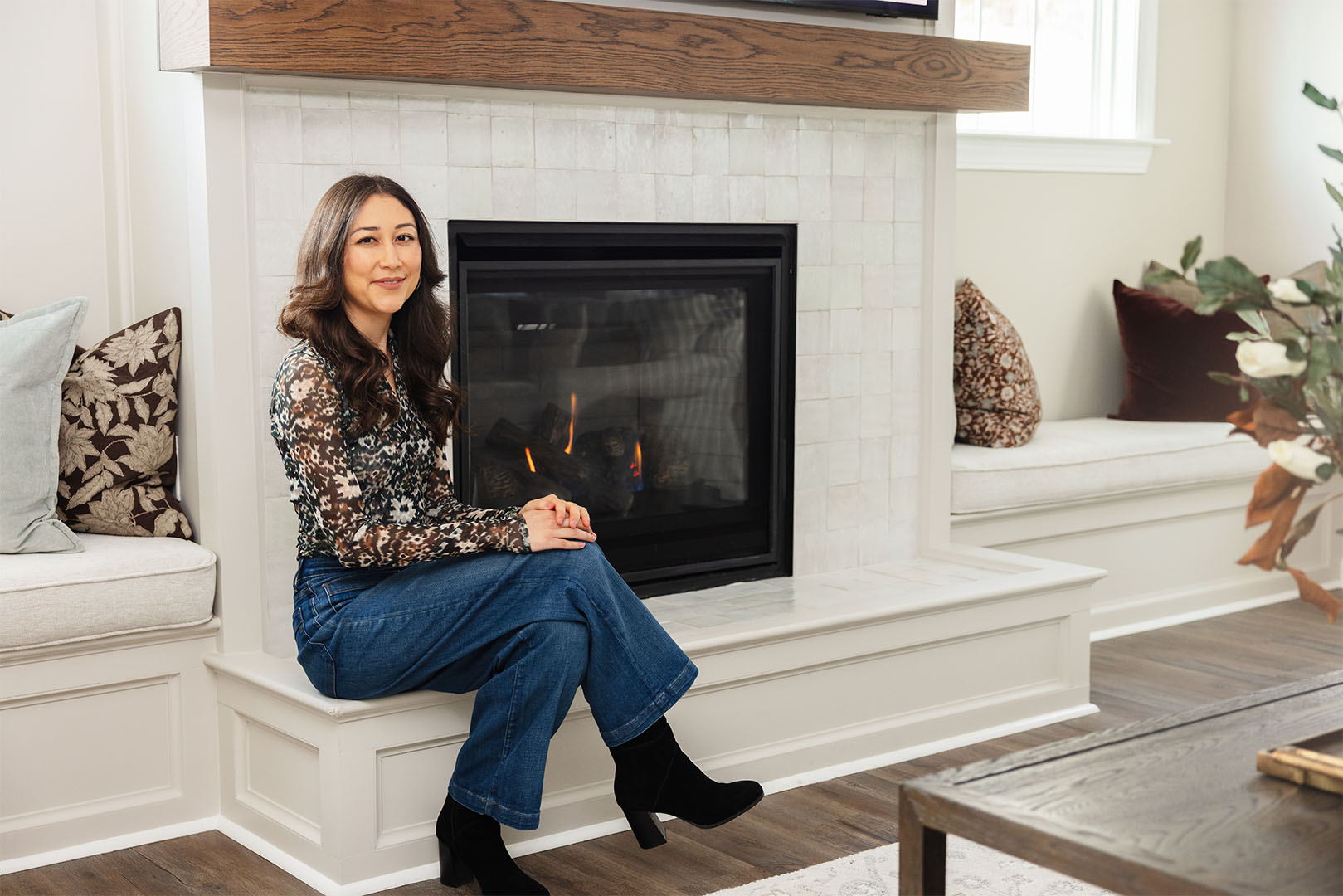 Woman with long dark hair sitting on a white fireplace ledge with a fire burning inside, smiling at the camera in a cozy living room with cushions.