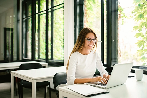 A woman is smiling as she sits at her laptop in an office.