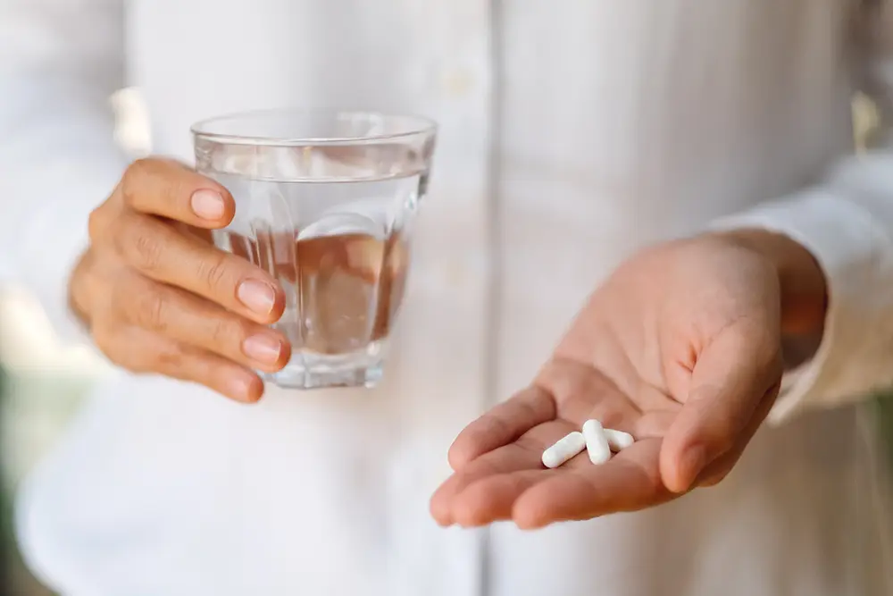 A person is holding a glass of water in one hand and tablets in the other.