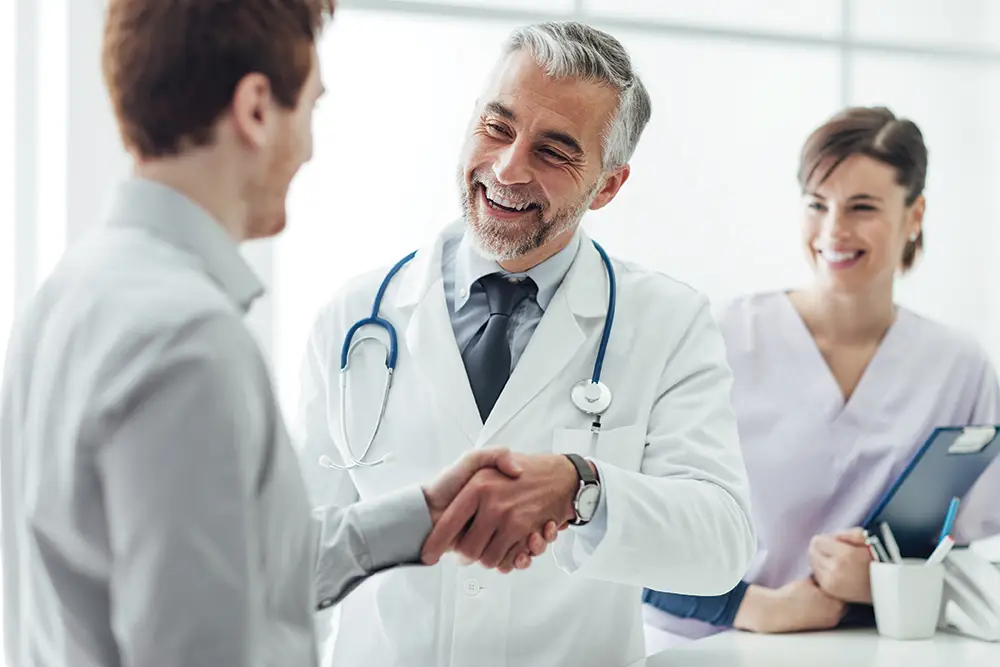 A doctor in a white coat shakes a patient's hand while a medical assistant stands nearby and they conduct a consultation.