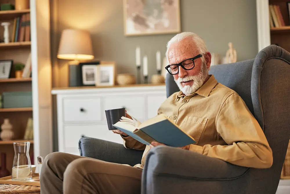 An older man sits happily in an armchair with a cup in his hand and reads a book.