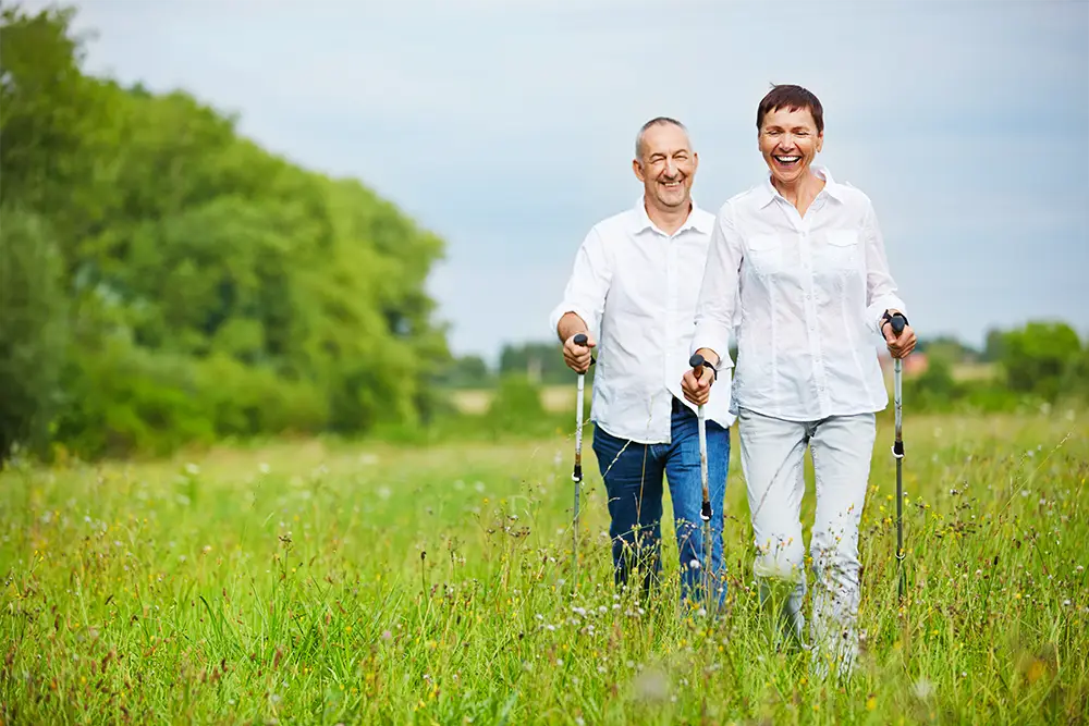 An older couple walk across a field with walking sticks.