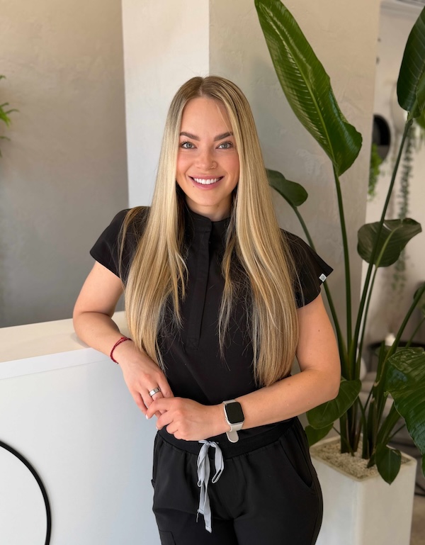 Usmívající se žena s dlouhými blond vlasy stojí u recepce s rostlinami v pozadí.