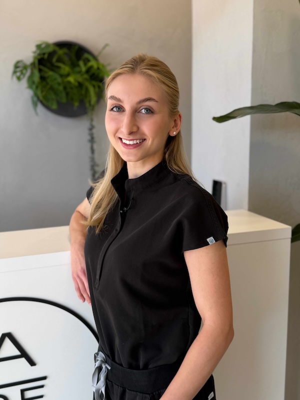 Usmívající se žena s blond vlasy v černém zdravotnickém oblečení opřená o recepci.