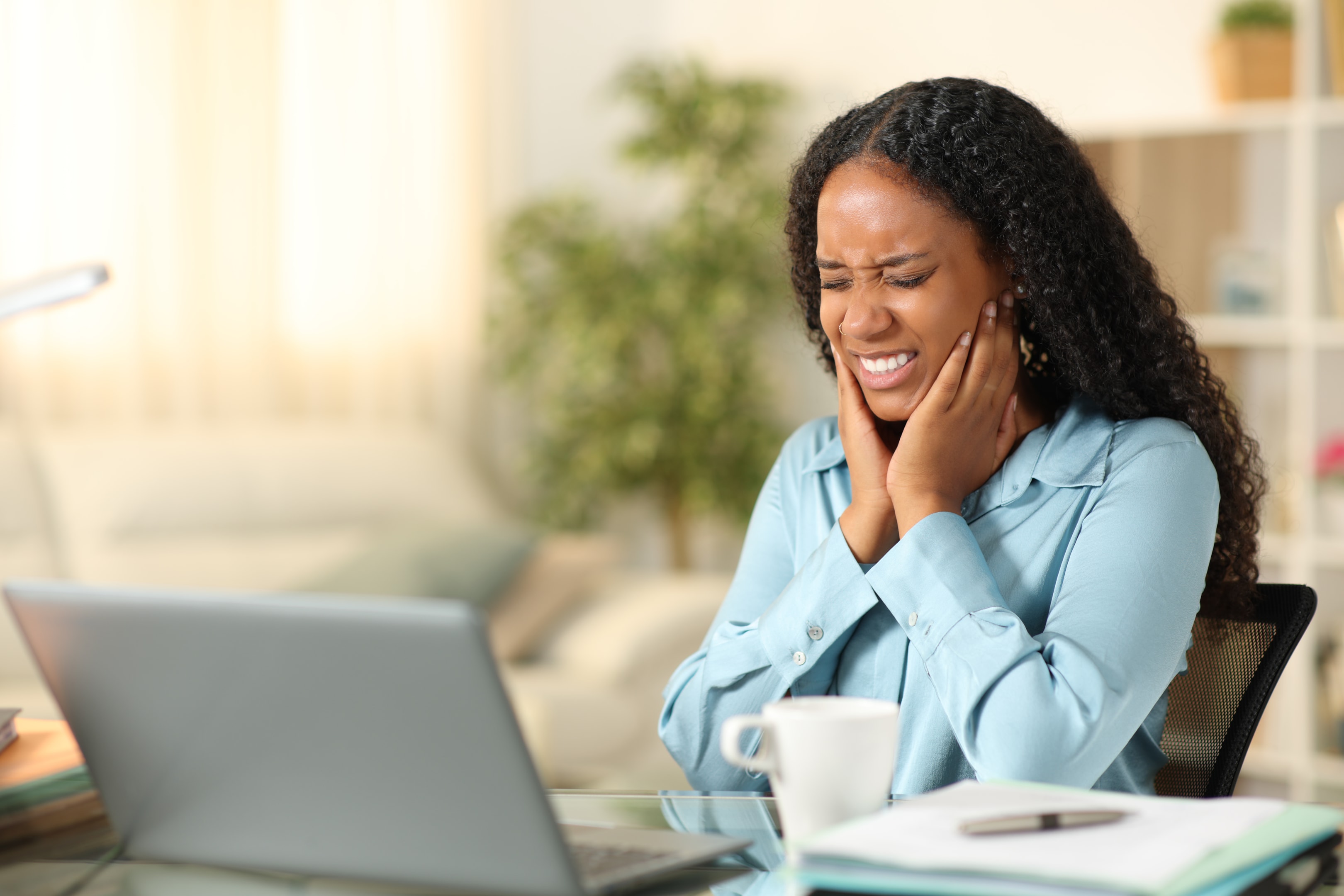 Woman in blue shirt holding her jaw in pain while sitting at desk with laptop in home office