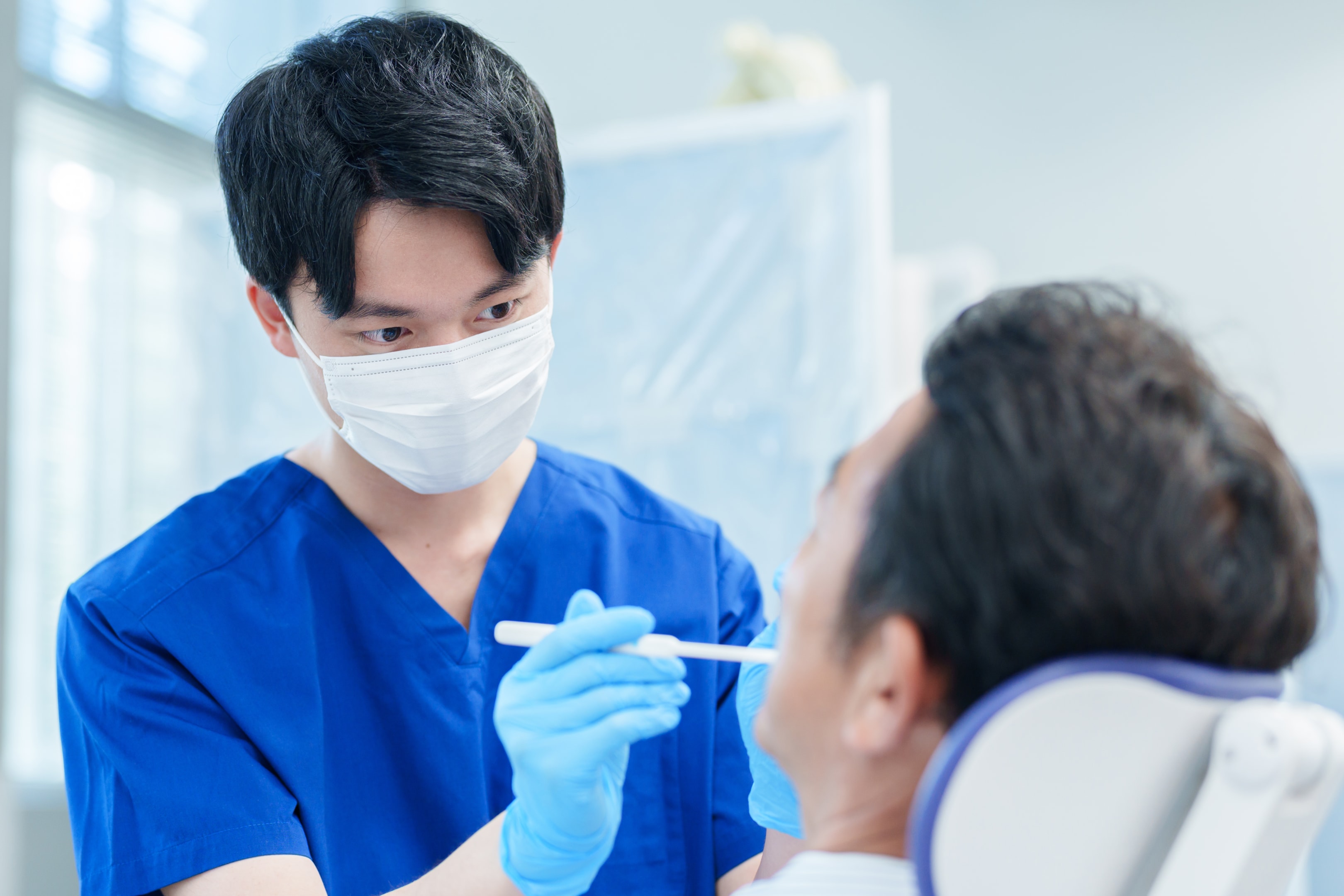 Healthcare worker in blue scrubs and mask examining patient with medical instrument in clinic.