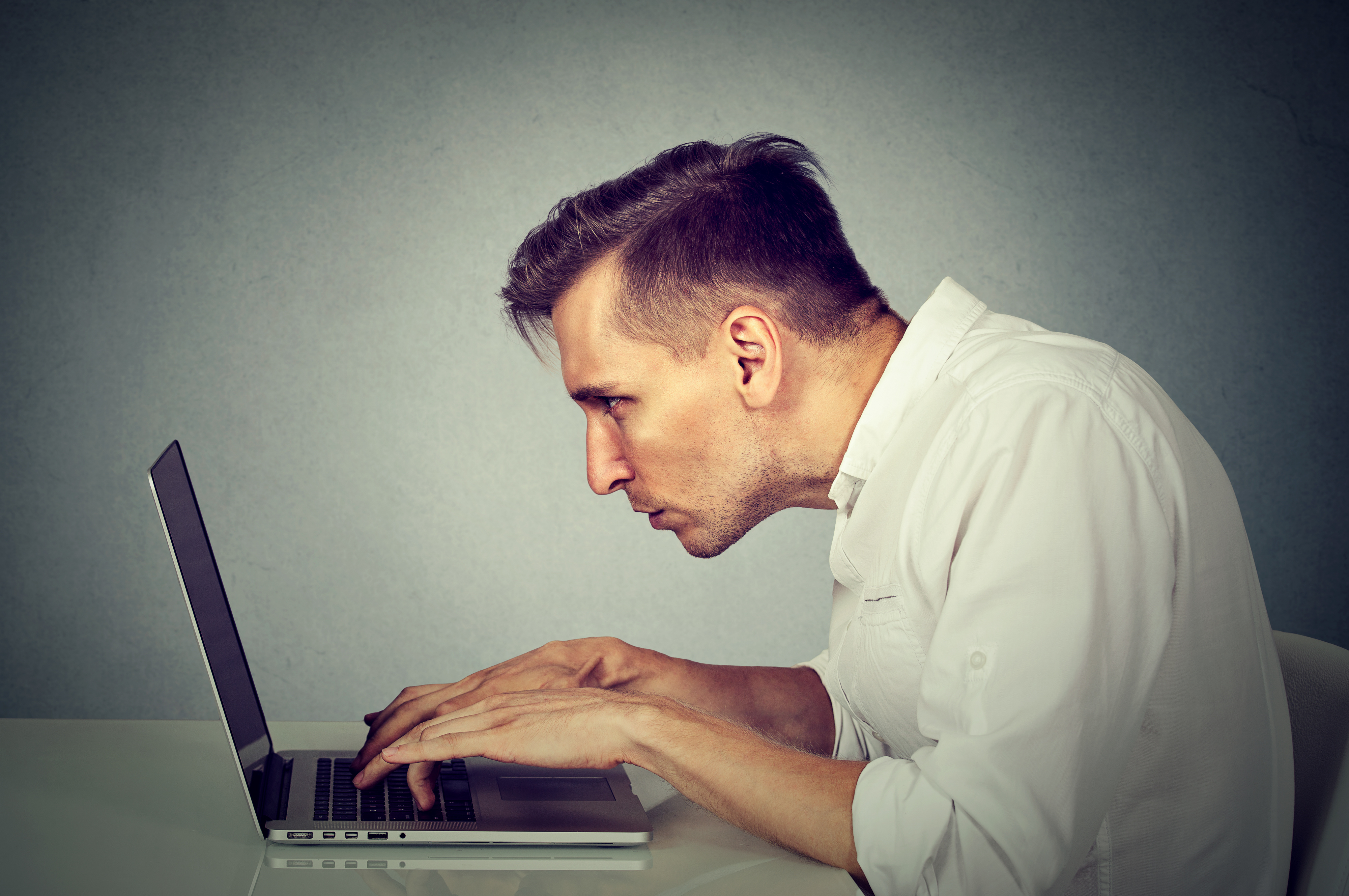 Man in white shirt hunched over laptop with poor posture, leaning close to screen while typing