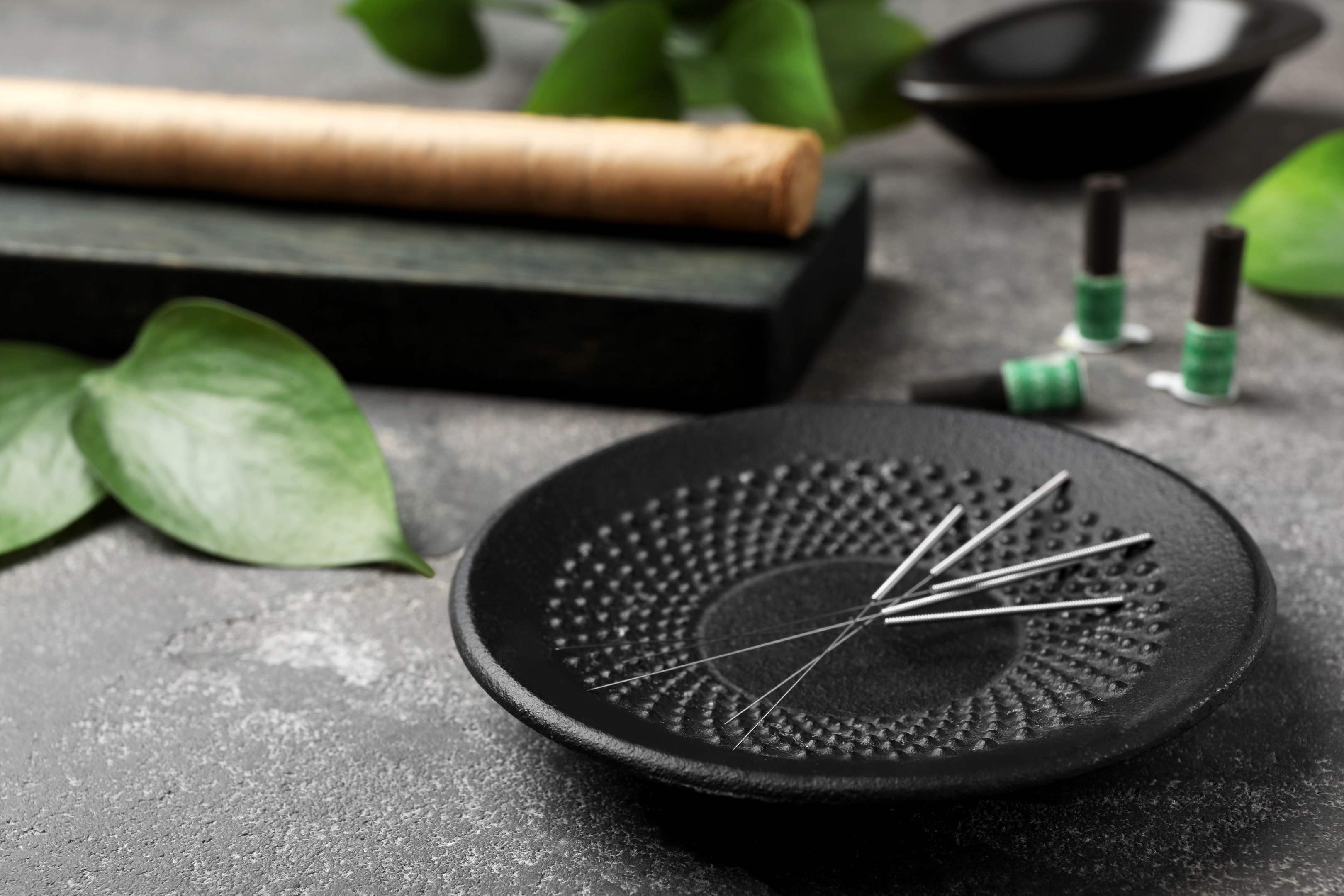 Black acupuncture plate with thin silver needles arranged on surface next to green plants and oils