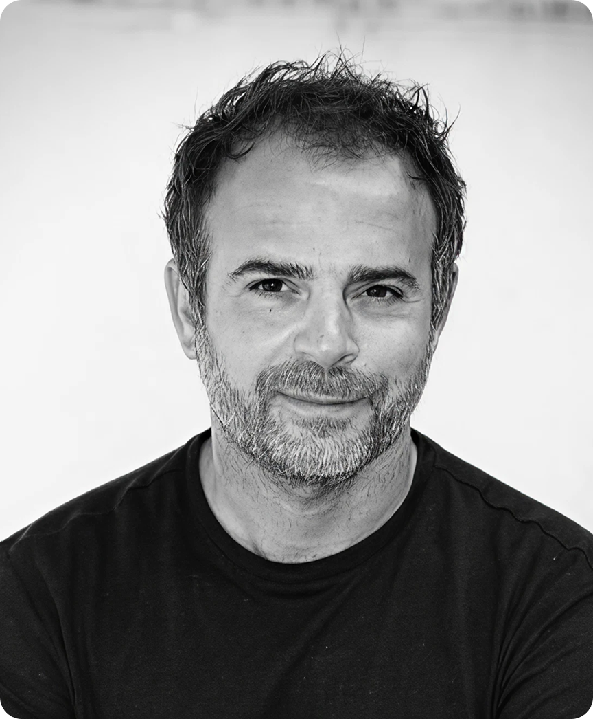 Portrait en noir et blanc d'un homme souriant avec barbe poivre et sel et cheveux en désordre, portant un t-shirt noir.