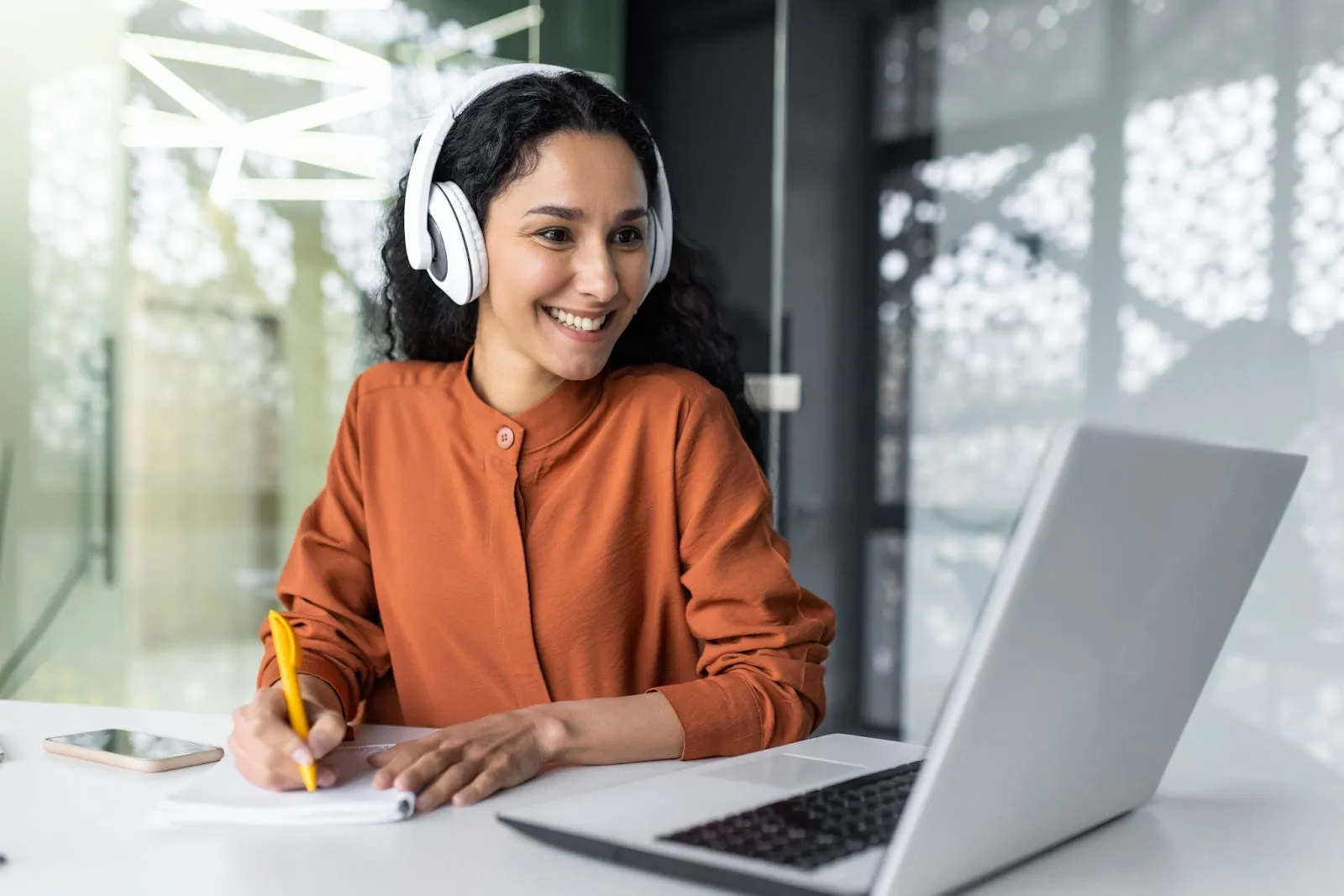Here is an alt text with dashes for the image you provided:smiling-woman-with-curly-hair-wearing-headphones-taking-notes-during-video-call-on-laptop-in-modern-office-setting-edge-tutor