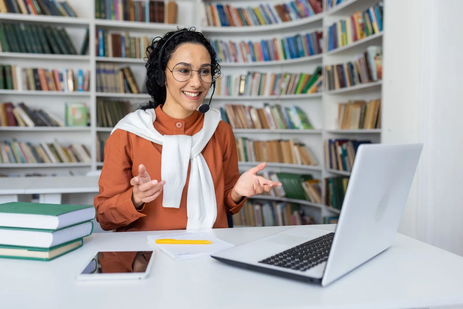 female-online-tutor-teaching-with-headset-in-library-smiling-at-laptop-with-books-tablet-and-notepad-on-desk-during-virtual-lesson-edge-tutor