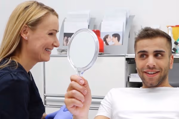Ein lächelnder Mann hält einen Handspiegel, während Dr. Berkei in blauen Handschuhen ihm in der Praxis gegenüber sitzt