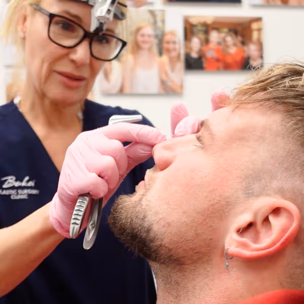 Plastische Chirurgin Dr. Berkei mit Brille und rosa Handschuhen untersucht die Nase eines Mannes in einer Klinik.