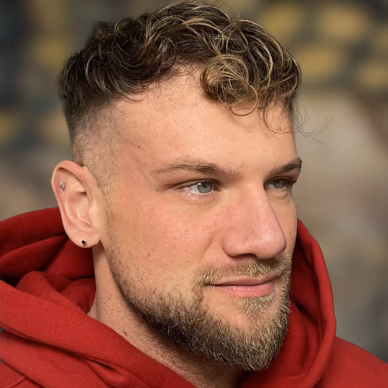 Porträt eines jungen Mannes mit kurzem lockigem Haar, Bart und rotem Kapuzenpullover, Patient in der Praxis von Dr. Berkei, blickt nach rechts.