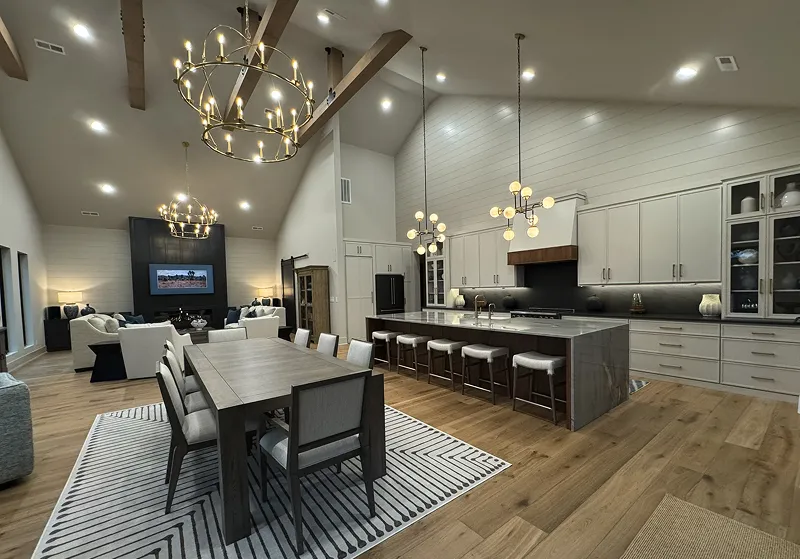 Spacious modern open-concept kitchen and living room with wooden floors, large dining table, kitchen island with stools, and multiple chandeliers.