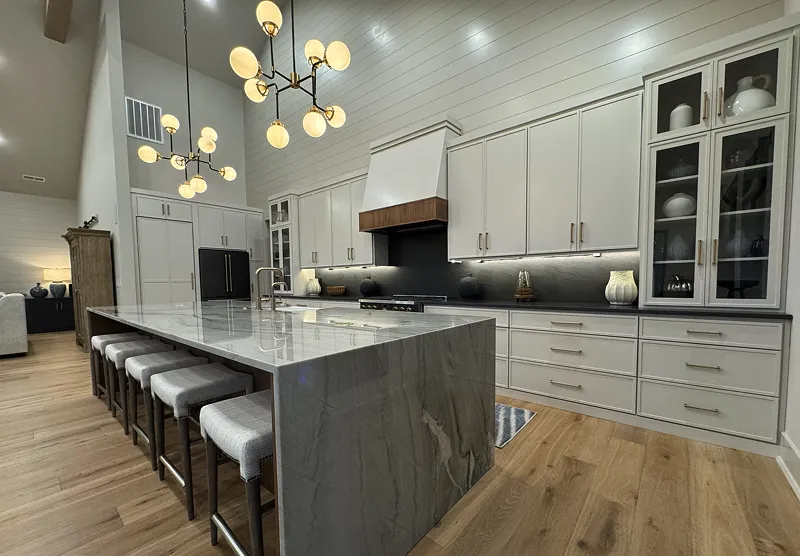 Modern kitchen with large marble island, six upholstered bar stools, white cabinets, black backsplash, wood accent range hood, and pendant lighting.