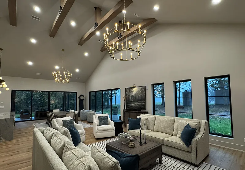 Spacious modern living room with high vaulted ceiling, exposed wooden beams, large windows, beige sofas, and elegant chandeliers.