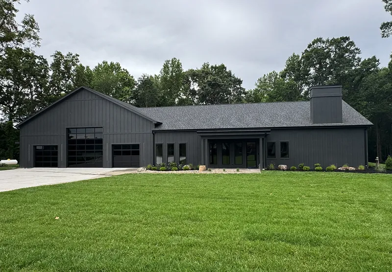 Modern single-story black house with large garage doors, surrounded by green lawn and trees under a cloudy sky.