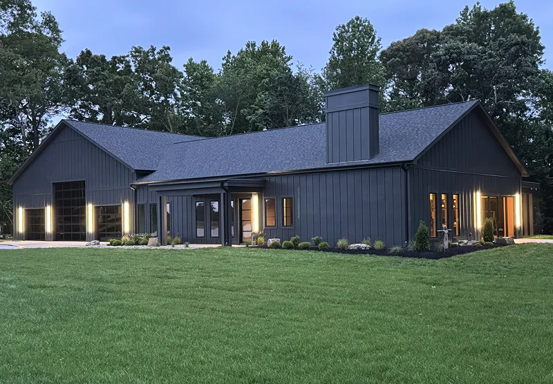 Modern dark gray barn-style house with large windows and warm interior lights, surrounded by green lawn and trees at dusk.