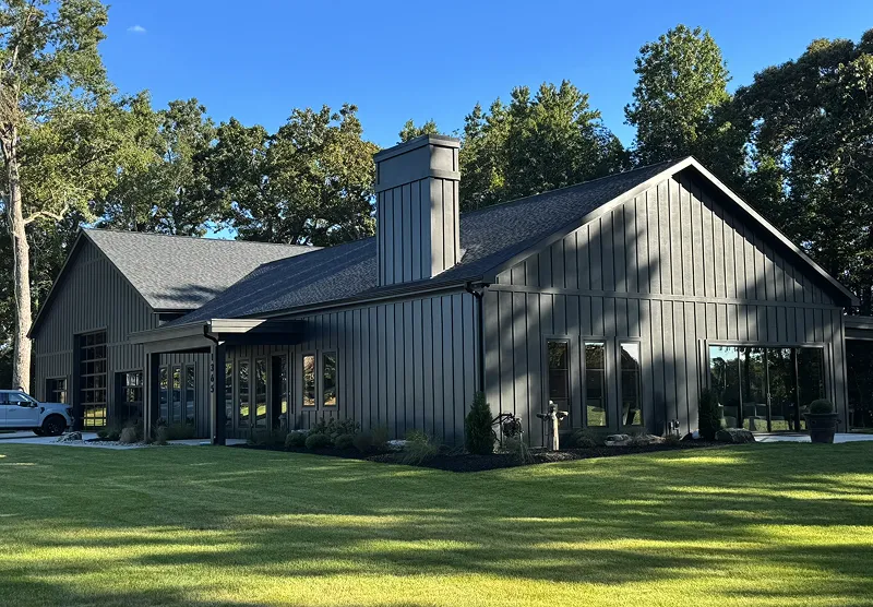 Modern gray house with vertical siding, large windows, a central chimney, surrounded by green lawn and trees under a clear blue sky.