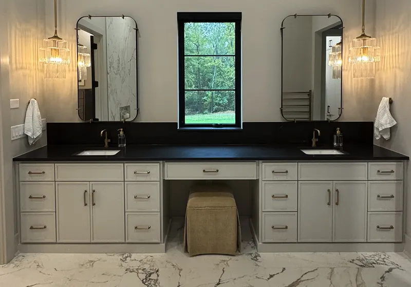 Modern double bathroom vanity with black countertop, two rectangular mirrors, hanging pendant lights, and a central cushioned stool under a middle window.