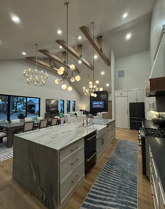 Modern open-concept kitchen and living room with a large marble island, pendant lighting, wood beams on the ceiling, a dining table, and large windows overlooking a backyard.