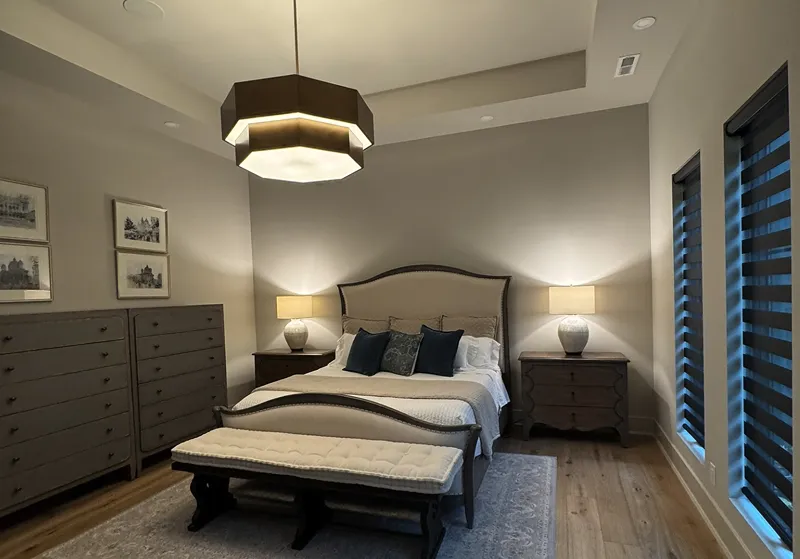 Modern bedroom with a beige upholstered bed, dark wooden nightstands with lamps, dressers, and a cushioned bench on a rug.