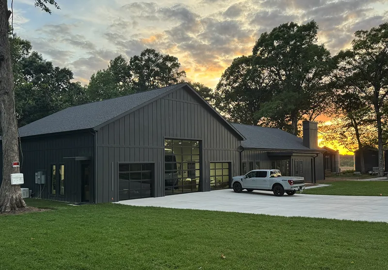 Modern dark gray barn-style building with large garage doors and a white pickup truck parked on a concrete driveway at sunset.