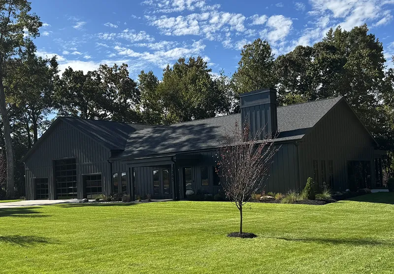 Modern dark gray single-story house with large glass garage doors, surrounded by green lawn and trees under a partly cloudy blue sky.