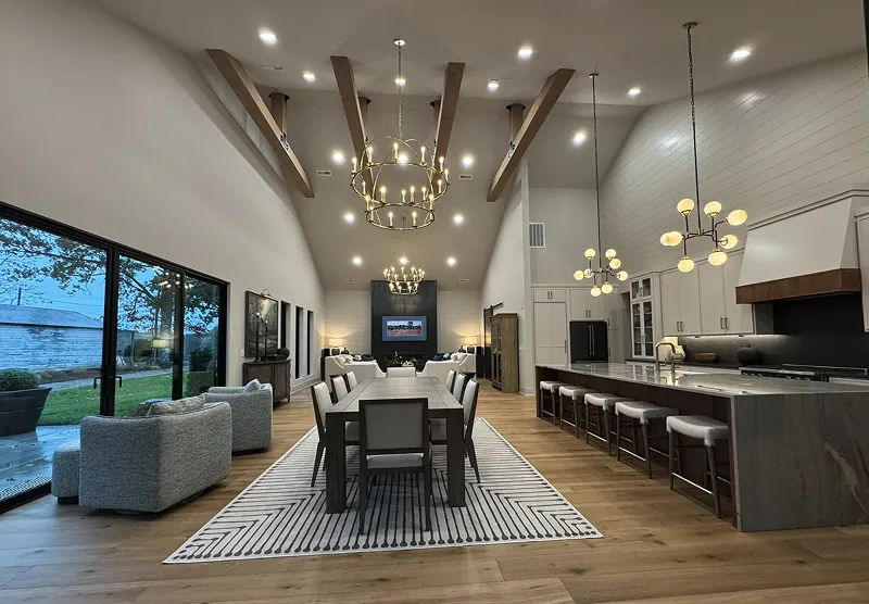 Spacious modern open-plan living area with a long dining table, multiple chandeliers, wooden ceiling beams, a large kitchen island with stools, and sliding glass doors to an outdoor patio.