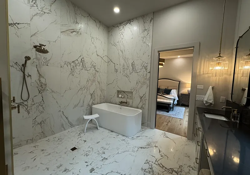 Modern bathroom with white marble walls and floor, freestanding bathtub, wall-mounted shower, and a vanity with black countertop next to an open door leading to a bedroom.