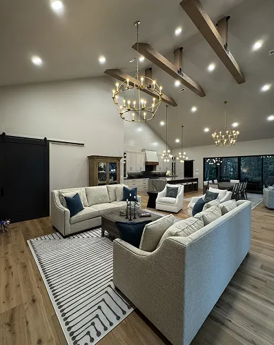 Modern open living room with high ceilings, wooden beams, gray sofas, striped rug, and large windows overlooking trees.