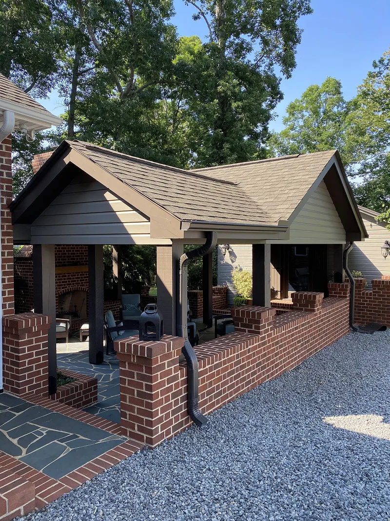 Outdoor covered patio with gray shingled roof, brick half wall, and stone flooring with chairs and a fireplace inside.