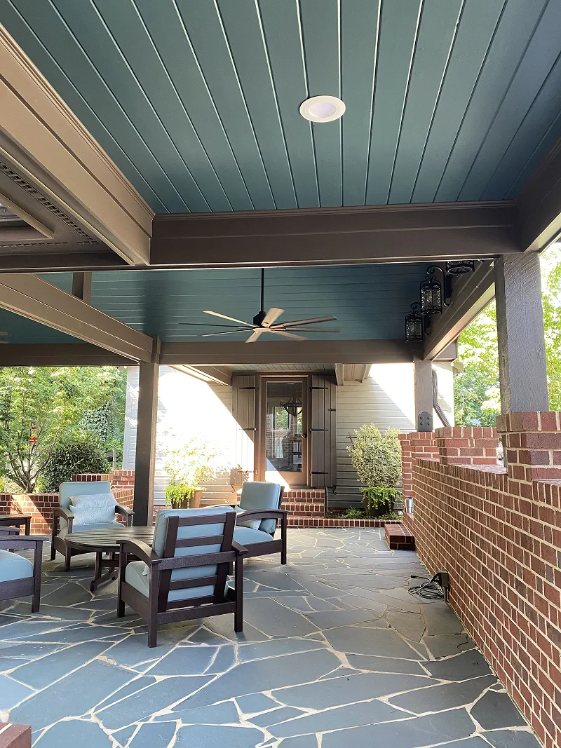 Covered outdoor patio with blue ceiling, ceiling fan, cushioned chairs around a wooden table, brick walls, and stone tile flooring.