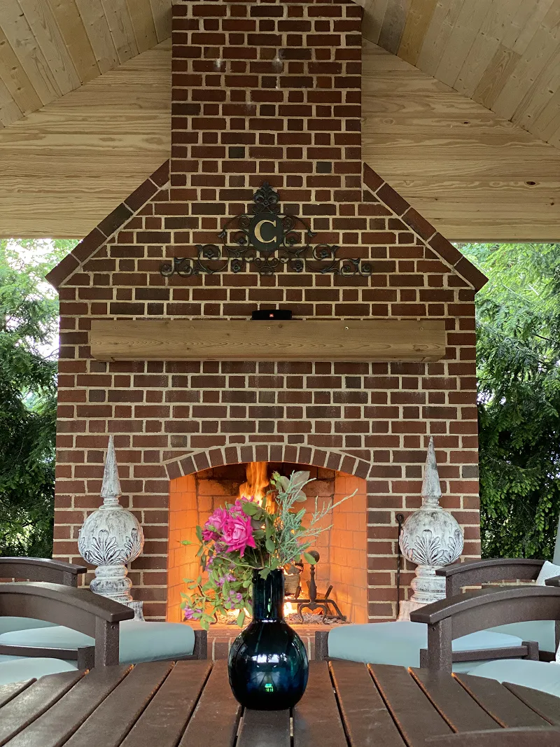 Outdoor brick fireplace with flames, decorative metal monogrammed letter C above mantel, and a vase with pink flowers on a wooden table in front.