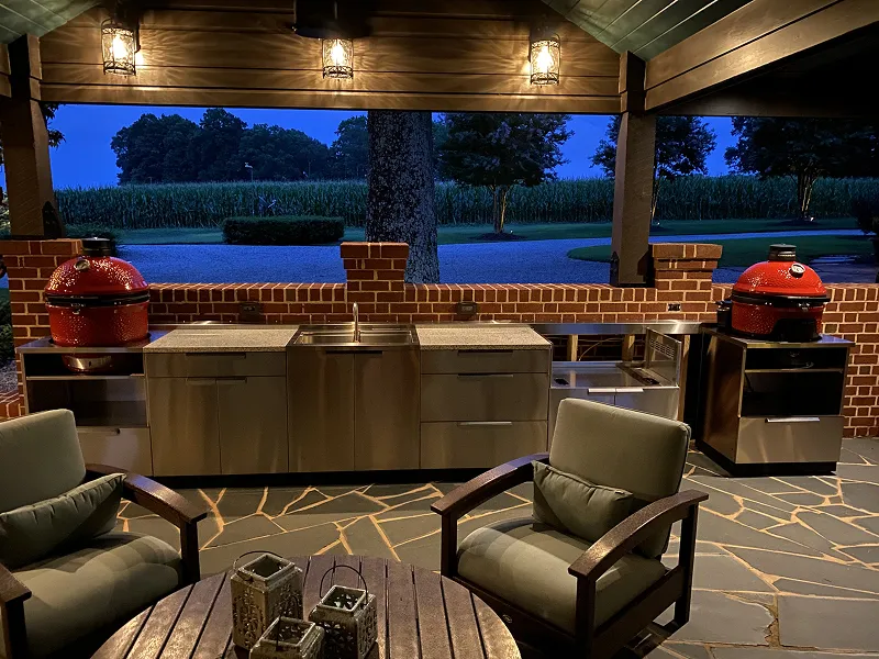 Outdoor covered kitchen at dusk with stainless steel cabinets, two red kamado grills, stone flooring, and two cushioned armchairs around a wooden table with lanterns.