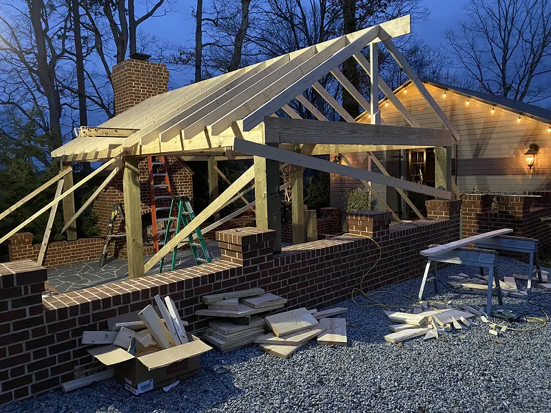 Wooden framework of a roof under construction over a brick patio area at dusk with ladders and scattered wood pieces nearby.
