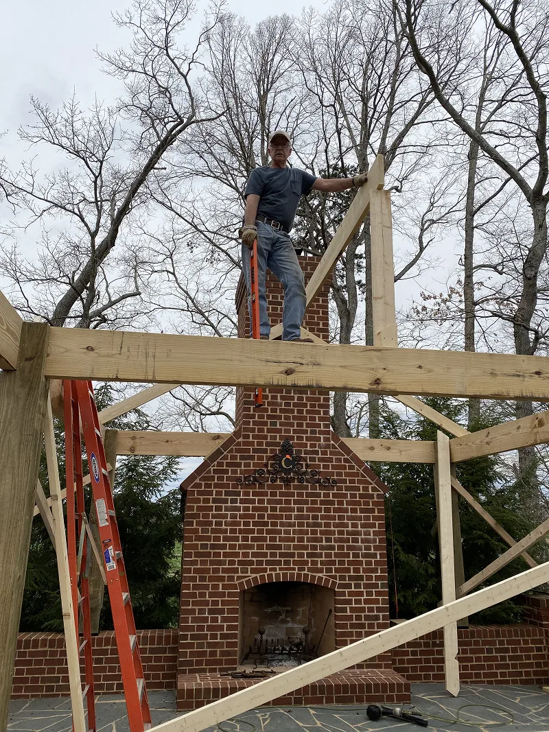 Man standing on wooden framework above a brick outdoor fireplace under construction in a backyard with leafless trees.