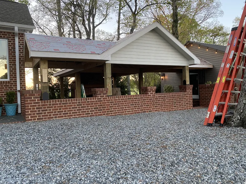 Partially constructed outdoor brick patio with wooden pillars and a gabled roof under construction, with a gravel ground and a red ladder nearby.