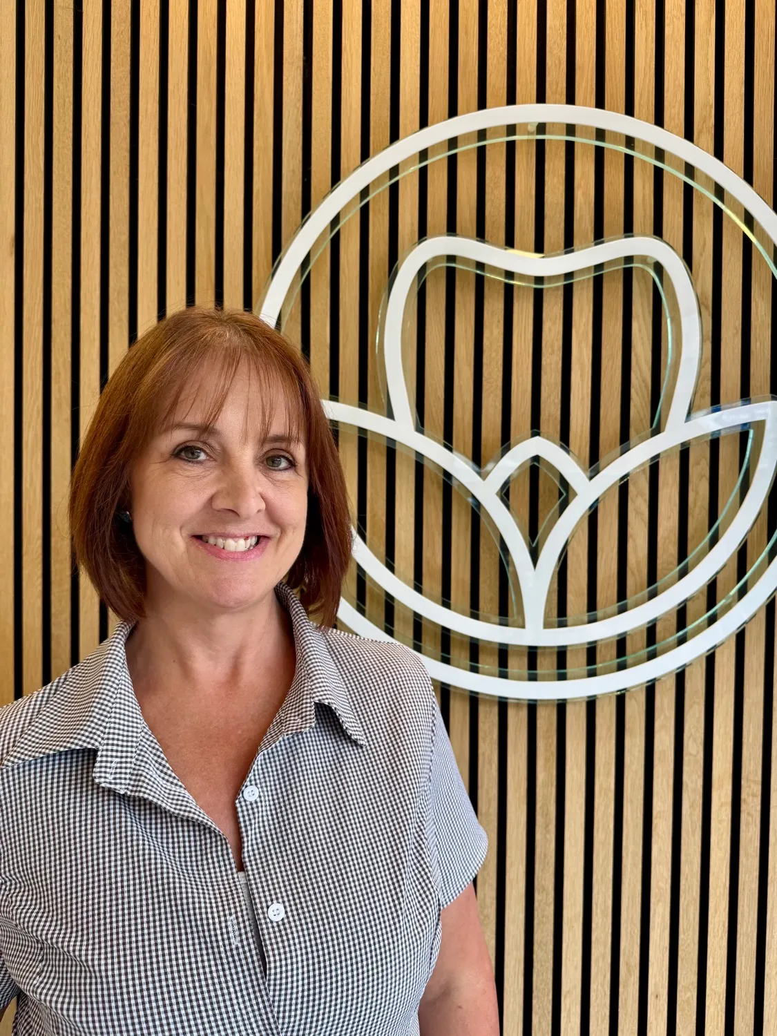 Smiling woman in checkered shirt in front of wooden wall and white logo
