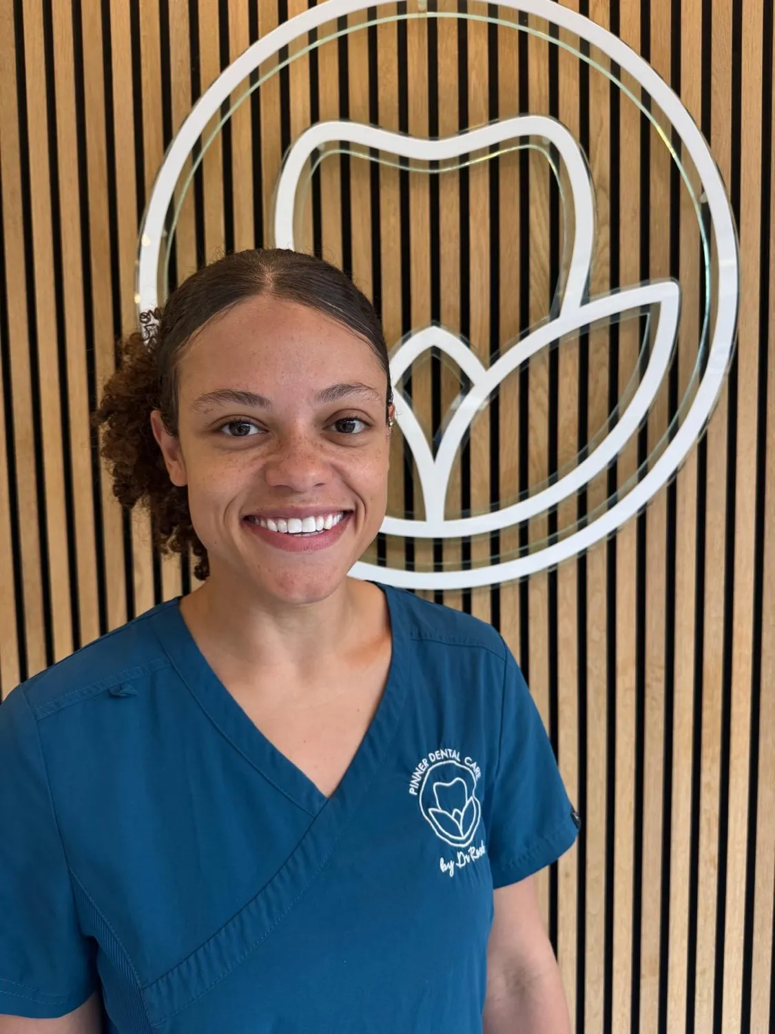 Smiling dental professional in blue scrubs standing in front of logo