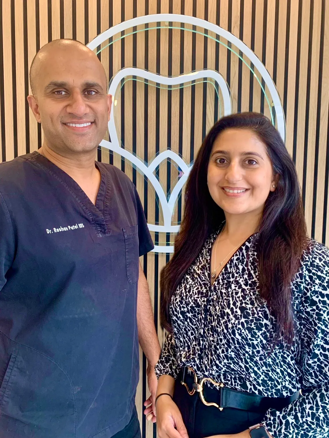 Smiling man wearing navy scrubs with embroidered name and woman in patterned blouse standing in front of a wooden wall with a large tooth emblem.