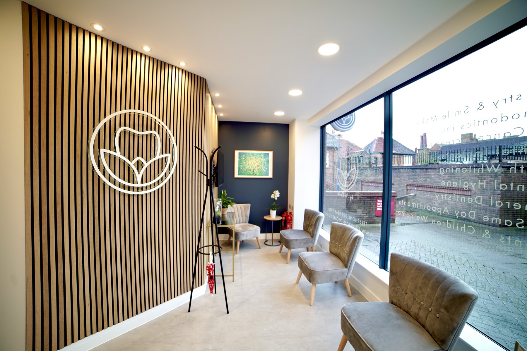 Bright waiting room with four gray velvet chairs, a wooden slatted wall with a white floral logo, large windows, and a coat rack.