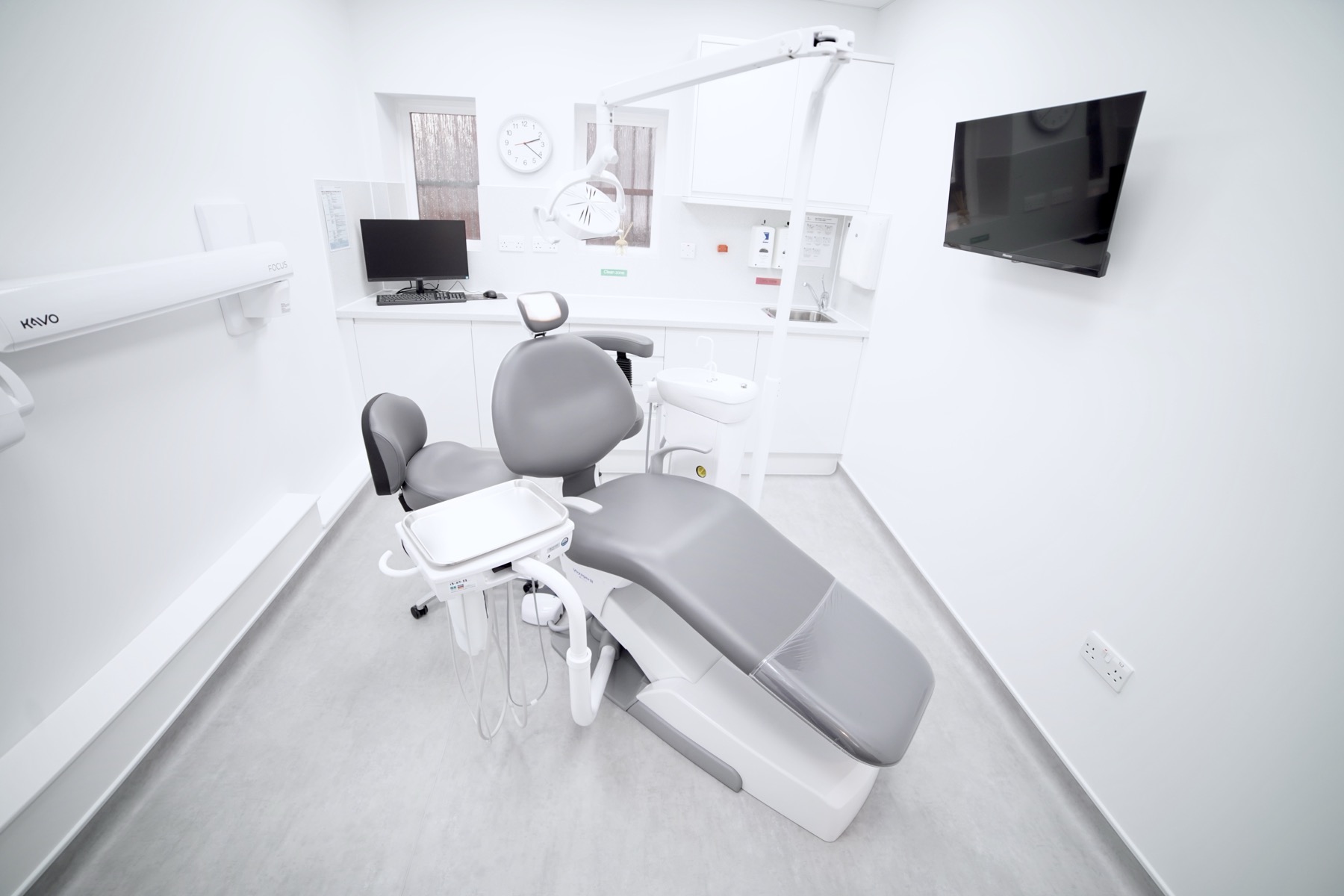 Modern dental clinic room with gray dental chair, overhead light, monitor, and clean white cabinetry.